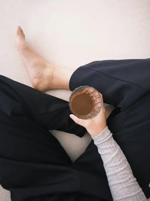 person wearing a gold anklet with black pants and a grey top on holding coffee