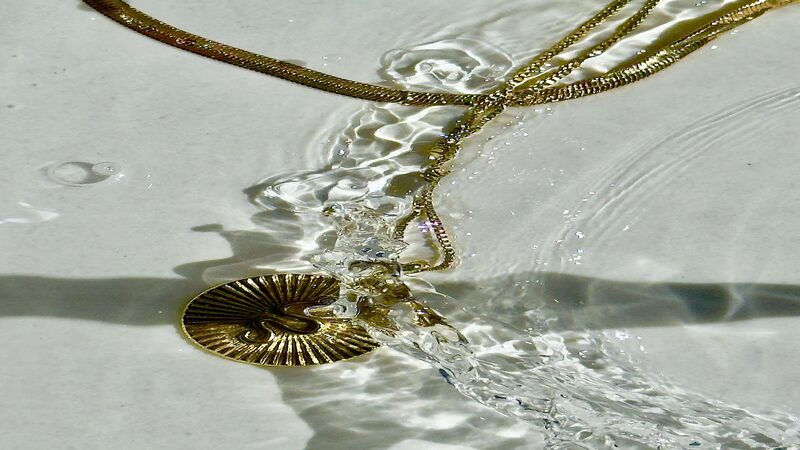 gold necklace with circle medal pendent and herringbone chain both submerged in water on a white background