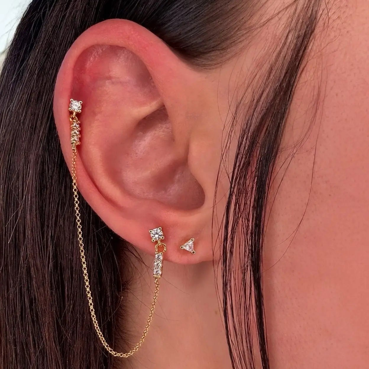 A person wearing a pair of gold-colored double stud earrings with a chain connecting the two studs, designed to mimic a single earring style.