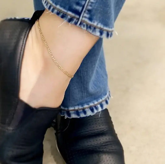 Close-up of a person wearing black shoes, blue jeans, and a gold anklet on a neutral background.