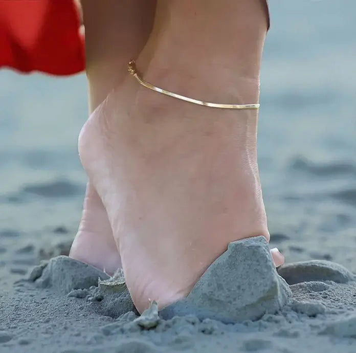 Close-up of a foot with a gold anklet on a sandy beach.