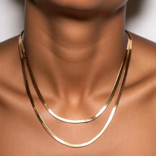 A close-up image of a woman's neck wearing a gold herringbone chain necklace.