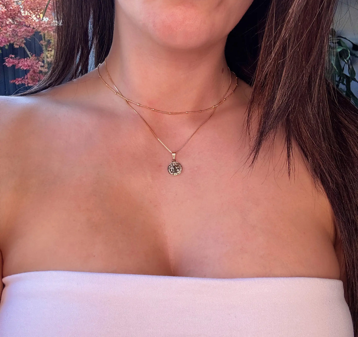 A close-up image of a person wearing a gold-colored pendant necklace with a textured small round charm, layered with a fine gold-filled chain.