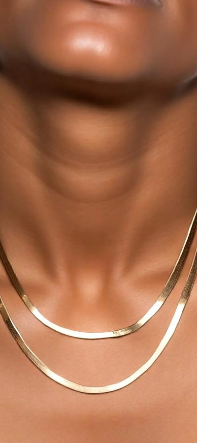 A close-up image of a woman's neck wearing a gold herringbone chain necklace.