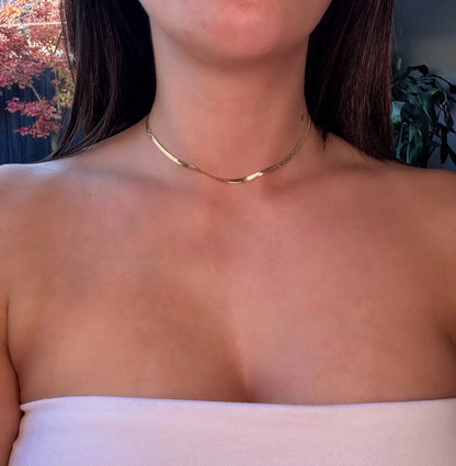 A close-up image of a woman's neck wearing a gold herringbone chain necklace.