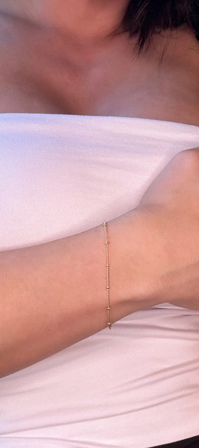 A close-up image of a person wearing a white strapless top, showcasing a gold-filled bracelet with small beads strung along a fine chain, resting on their wrist.