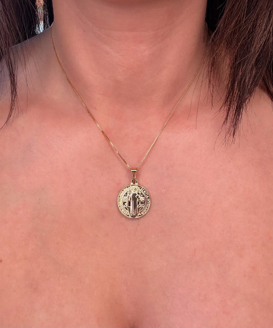 A close-up image of a person wearing a gold-colored pendant necklace with a textured large round charm, layered with a fine gold-filled chain.