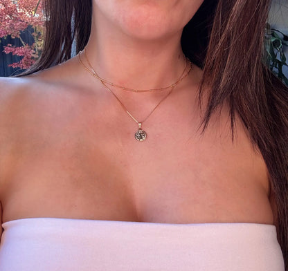 A close-up image of a person wearing a gold-colored pendant necklace with a textured small round charm, layered with a fine gold-filled chain.